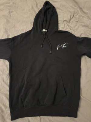 Svart hoodie från Your Turn - Svart hoodie från Your Turn med vit logga på bröstet och stor text på ryggen. Klassisk känguruficka framtill och justerbar huva med snören. Tillverkad i mjukt bomullsmaterial som är skönt att bära. Perfekt för en avslappnad och trendig stil.