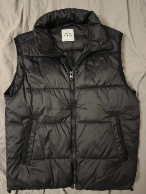 Svart dunväst från ZARA - Stilren svart dunväst från ZARA med hög krage och dragkedja framtill. Västen har två snedställda fickor med dragkedja och quiltad design. Perfekt för lager-på-lager och enkel att matcha med olika outfits. Finns lite slitache på baksidan se sista bilden.