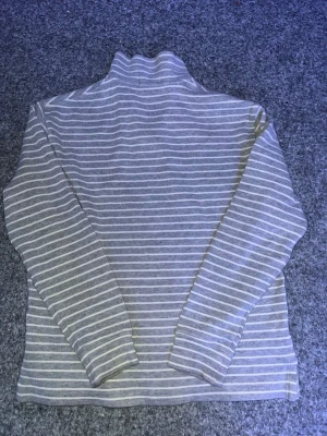 Gråvit randig tröja från Polo Ralph Lauren - Säljer en grå och vit randig tröja från Polo Ralph Lauren med halv dragkedja och hög krage. Tröjan har långa ärmar och den klassiska Polo-loggan broderad på bröstet. Perfekt för dig som gillar stilrena och bekväma plagg.