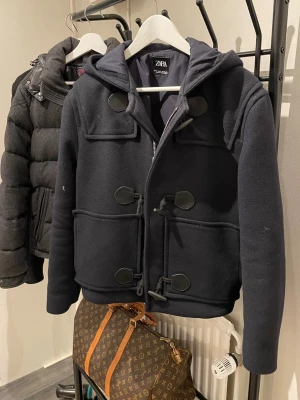 Zara jacka - Stilren marinblå Zara jackamed huva och klassiska knäppningar i träimitation men även med dragkedja. Perfekt för kyliga dagar och enkel att matcha med olika outfits. Batalade 1499 kr för den och säljer den för 900 kr (Bild med den på kan visas i dm)‼️