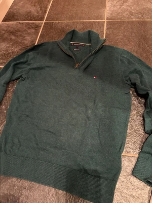 Grön halvzip-tröja Tommy Hilfiger - Mörkgrön stickad tröja från Tommy Hilfiger med halv dragkedja och liten broderad logga på bröstet. Tröjan är i bomull och har ribbade muddar vid ärmslut och nederkant. Perfekt för lager-på-lager och snygg till jeans.