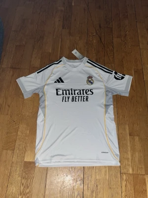 Real Madrid matchtröja Adidas vit S - Säljer en vit Real Madrid matchtröja från Adidas med svarta detaljer och gula inslag. Tröjan har klubbmärke och sponsortryck på bröstet samt svarta ränder på axlarna. Tillverkad i lätt och ventilerande material, perfekt för fotboll eller träning.
