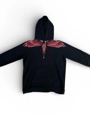 Marcelo Burlon - Säljer en svart hoodie från Marcelo Burlon med ikoniskt rött vingtryck över axlar och bröst, samt på ryggen. Hoodien har huva och en stor magficka framtill. Tillverkad i mjukt bomullsmaterial och har en avslappnad passform. Perfekt för dig som gillar streetwear och unika detaljer.
