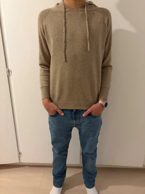Beige hoodie i mjukt material - Säljer en stilren beige hoodie med huva och dragsnören. Hoodien har långa ärmar, ribbade muddar och är gjord i ett mjukt, tunt stickat material som känns skönt mot huden. Perfekt för dig som gillar en enkel och clean look. Modellen i bilden är 170 och den sitter bra.