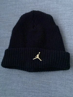 Svart ribbstickad Jordan mössa - Säljer en svart ribbstickad mössa från Jordan med uppvikt kant och guldfärgad Jumpman-logga framtill. Klassisk och stilren design som passar perfekt till streetwear och vardagsoutfits.