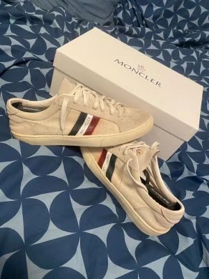 Beige Moncler sneakers  - Snygga beige sneakers från Moncler med klassiska röda och blå ränder på sidan samt Moncler-logga. Skorna har lågt skaft, vita snören och är tillverkade i mocka. Perfekta för dig som vill ha en stilren och exklusiv look.