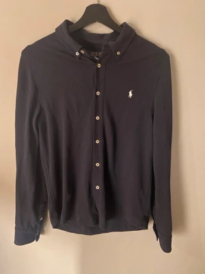 Mörkblå skjorta från Polo Ralph Lauren - Klassisk mörkblå skjorta från Polo Ralph Lauren med vit broderad logga på bröstet. Skjortan har långa ärmar, knappar framtill och en mjuk krage. Perfekt för en stilren och avslappnad look. Storlek L i barnavdelning. Passar XS