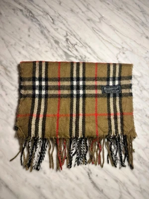 beige halsduk Burberry - Snygg beige halsduk från Burberry med ikoniskt rutmönster i svart, vitt och rött. Halsduken har fransar i ändarna och en vävd Burberry-etikett. Perfekt accessoar för att lyfta din outfit med en tidlös brittisk touch.