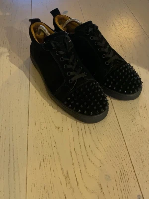 Svarta sneakers med nitar Christian Louboutin - Säljer ett par svarta sneakers från Christian Louboutin med coola svarta nitar på tån och klassisk röd sula. Jag har personligen storlek 43 i skor men de passar. Skorna har svarta snören, detaljer i mocka och skinn samt guldfärgad insida. Perfekta för dig som vill sticka ut med din stil.
