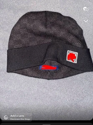 Svart ribbad mössa med patch - Säljer en svart ribbad mössa med uppvikt kant och en fyrkantig patch framtill. Mössan har diskret rutmönster i materialet och en klassisk form som passar till det mesta. Perfekt för dig som vill ha en stilren accessoar till hösten och vintern.