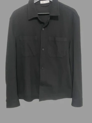 Svart skjorta från Zara med fickor - Stilren svart skjorta från Zara med två bröstfickor och knäppning framtill. Skjortan har klassisk krage och långa ärmar. Perfekt för dig som gillar en enkel men snygg look.