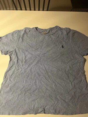 Ljusblå t-shirt från Polo Ralph Lauren - Ljusblå t-shirt från Polo Ralph Lauren med klassisk rund hals och liten broderad logga på bröstet. Tillverkad i mjuk bomull och har en avslappnad passform. Perfekt för en stilren och enkel look.