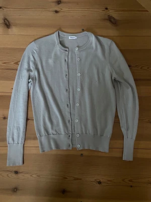 Ljusgrå cardigan från Filippa K - Snygg ljusgrå cardigan från Filippa K i tunn, mjuk bomull. Klassisk rund halsringning och knäppning med vita knappar framtill. Långa ärmar och ribbade muddar ger en clean och stilren look. Perfekt att slänga över en t-shirt eller linne.