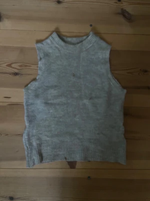 Beige stickad väst med ribbad kant - En enkel och stilren beige stickad väst utan ärmar. Västen har en hög, rund halsringning och bred ribbad kant nedtill. Perfekt att bära över en skjorta eller t-shirt för en avslappnad men trendig look.