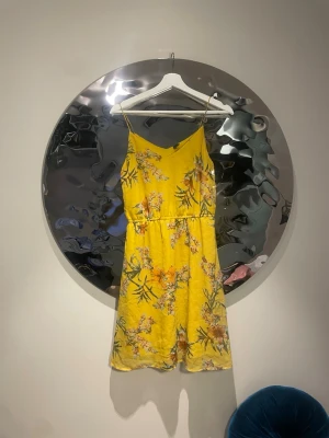 Gul blommig sommarklänning - Säljer en gul kortklänning med blommigt mönster och smala axelband. Klänningen har en v-ringad hals och är tillverkad i ett lätt material, perfekt för varma dagar. Passformen är normal och klänningen har en avslappnad, luftig känsla.