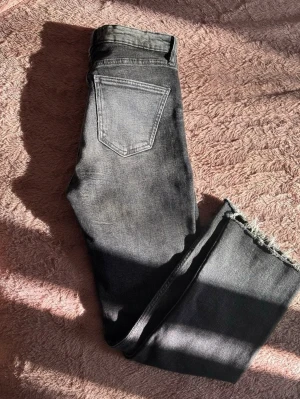 Svarta raka jeans från Zara - Säljer ett par svarta jeans från Zara med raka ben, movie washed look och råa, fransiga benslut. Klassisk femficksmodell och hög midja. Perfekta till en avslappnad och trendig stil. Materialet är bomull med lite stretch för extra komfort.