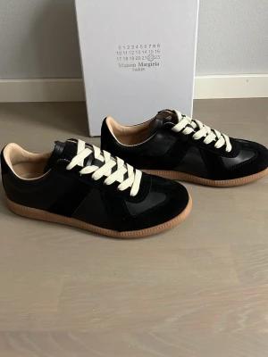 Maison Margiela svarta sneakers - Säljer ett par snygga Maison Margiela sneakers. Skorna är svarta med vita snören och beige sula. Storlek 42🙌box och dustbag finns. Tveka inte att höra av er vid frågor🤝pris är såklart diskuterbart! 💸