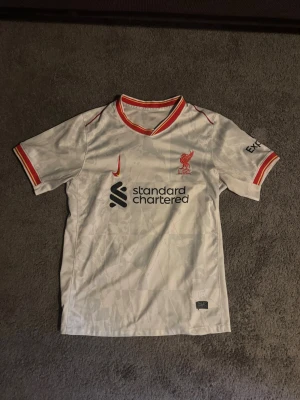 Vit Liverpool matchtröja Nike - Säljer en vit Liverpool matchtröja från Nike med röda och gula detaljer på krage och ärmslut. Tröjan har Liverpool FC-logga broderad på bröstet, Nike-logga och sponsortryck framtill. Tillverkad i lätt och ventilerande material, perfekt för fotbollsträning.