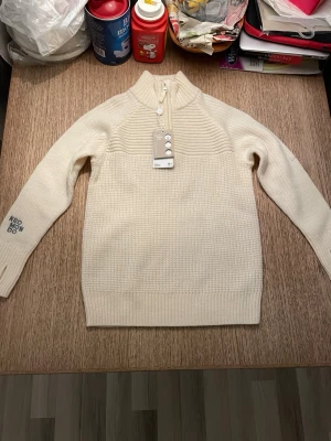 Vit stickad tröja med halv dragkedja från Neomondo - Snygg krämvit stickad tröja från Neomondo med halv dragkedja och hög krage. Tröjan har ribbade muddar och texten 'NEOMONDO' på ena ärmen. Perfekt för lager på lager och för att hålla sig varm.