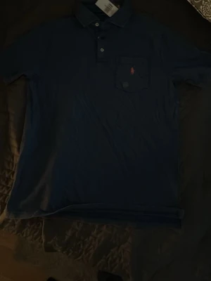 Blå pikétröja från Polo Ralph Lauren - Säljer en klassisk blå pikétröja från Polo Ralph Lauren i bomull. Tröjan har krage, knappar framtill och en liten ficka på bröstet med den ikoniska loggan broderad. Classic fit-modell som passar till det mesta.