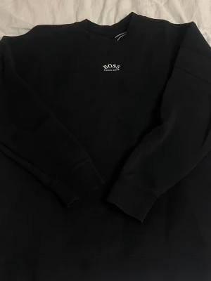 Svart sweatshirt från Hugo Boss - Svart sweatshirt från Hugo Boss med liten logga framtill och stor BOSS- och HUGO BOSS-tryck på ryggen. Klassisk rund halsringning och ribbade muddar vid ärmslut och nederkant. Perfekt för en clean och stilren look.