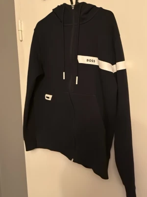 Svart hoodie med vit detalj från France BOSS - Snygg svart hoodie från BOSS med vit rand och logga på bröstet. Hoodien har dragkedja, justerbar huva med snören och en stor magficka. Tillverkad i mjukt material som känns skönt mot huden. Perfekt för en avslappnad och trendig look.