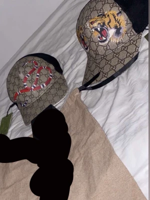 Gucci keps med tiger och orm - Två kepsar från Gucci med klassiskt GG-mönster. Ena kepsen har en färgglad orm på framsidan, den andra har en tiger med öppet gap. Båda har svart mesh på baksidan och justerbar rem. Perfekt för dig som vill sticka ut med lyxiga accessoarer även äkta ifrån Italien gucci.