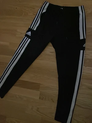 Svarta Adidas leggings med vita ränder - Säljer ett par svarta Adidas leggings med de klassiska tre vita ränderna längs benen och Adidas-logga på låret. Tight passform och dragsko i midjan. Perfekta för träning eller chill. Materialet är mjukt och stretchigt, troligtvis polyester och elastan.