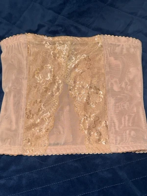 Beige korset - Säljer ett beige korse med vackra spetsdetaljer och blommönster framtill. Plagget är formande och har en bred, elastisk design som ger stöd runt midjan. Materialet är mjukt och stretchigt, perfekt för att skapa en slät siluett under kläder.