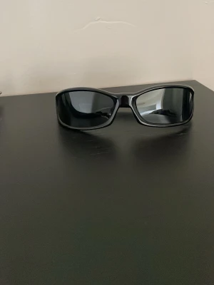 Svarta sportiga solglasögon - Säljer ett par svarta solglasögon med sportig design och breda bågar. Glasen är mörka och bågarna är tillverkade i syntetmaterial. Perfekta för dig som vill ha en clean och stilren look.
