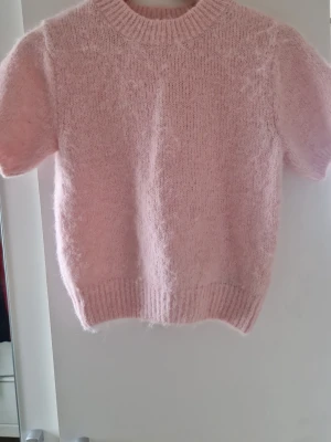 Ljusrosa stickad tröja Gina Tricot - Supersöt ljusrosa stickad tröja från Gina Tricot med korta ärmar och ribbade muddar vid hals, ärmslut och nederkant. Tröjan har en fluffig och mjuk känsla, perfekt för att addera en gullig touch till din outfit.