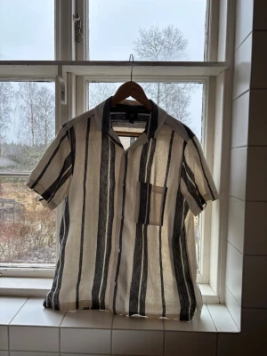 Randig kortärmad linneskjorta - Snygg kortärmad skjorta i linne med vertikala svarta och vita ränder. Skjortan har öppen krage och en bröstficka på vänster sida. Skön inför sommaren och våren då den funkar som overahirt också. Den ser lite  beige/gul ut på bilderna men är egentligen vit. Storlek S men passar även de som ligger mellan S och M. 