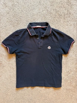 Mörkblå Moncler pikétröja - Snygg mörkblå pikétröja från Moncler med klassisk krage och knappar framtill. Tröjan har röda och vita detaljer på krage och ärmslut samt Moncler-logga på bröstet. Perfekt till jeans eller kjol för en sportig look.