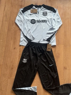 Nike FC Barcelona träningsset - Snyggt träningsset från Nike och FC Barcelona med långärmad, ljusgrå tröja med svart axelparti och halv dragkedja. Tröjan har FCB-logga och Spotify-tryck. Matchande svarta byxor med Nike-logga och FCB-märke. Perfekt för fotbollsträning.