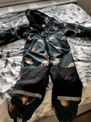 Gr Spårdräktgle täckbyxa Minimarket - Mörkgrön täckbyxa/overall från Minimarket med huva och gulligt lejonmönster. Förstärkta svarta partier på knän och459 ben, samt reflexdetaljer. Perfekt för kalla dagar och lek utomhus. Passar barn 6-7 år NB! Säljs utan accessoarer.