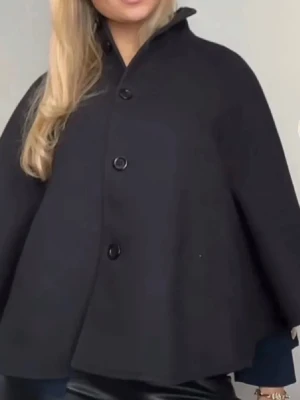 Svart poncho kappa  - Stilren svart cape-kappa med tre stora knappar framtill och hög krage. Kappan har en vid, svepande passform och är tillverkad i ett tjockare tyg som ger en elegant siluett. Perfekt för dig som vill ha en trendig och unik ytterjacka.