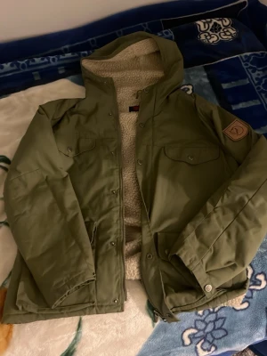 Grön teddyjacka från Fjällräven - Säljer en grön teddyfodrad jacka från Fjällräven med huva och tryckknappar. Jackan har två bröstfickor med lock och ett mjukt, beige teddyfoder på insidan. Perfekt för kyliga dagar och har en klassisk outdoor-look.