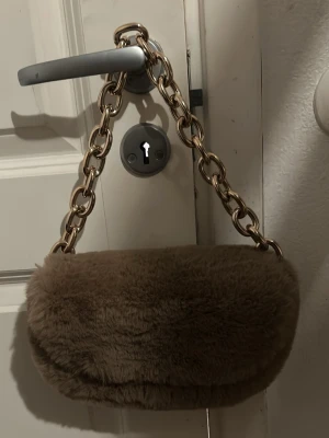 Beige fluffig handväska med kedja - Supermjuk och fluffig beige handväska med stor guldfärgad kedja som axelrem. Väskan har ett foder i matchande beige tyg och ett litet innerfack. Perfekt för dig som vill ha en trendig accessoar med mysig känsla.