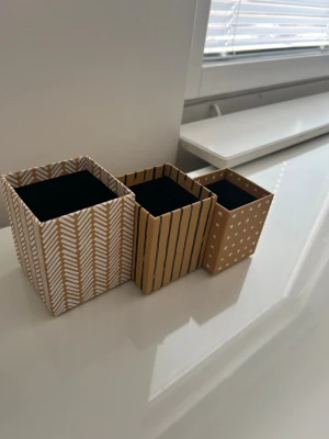 Tre dekorativa förvaringslådor - Säljer tre fyrkantiga förvaringslådor i olika storlekar. Lådorna är i beige och vitt med olika mönster: prickar, ränder och pilmönster. Perfekta för att organisera småsaker på skrivbordet eller i hemmet. Tillverkade i hård papp eller tunn kartong.