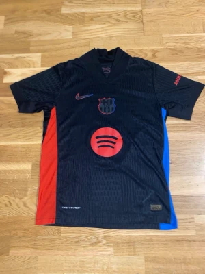 FC Barcelona Lamine Yamal matchtröja - Snygg svart FC Barcelona matchtröja från Nike med Lamine Yamal och nummer 19 på ryggen. Tröjan har röda och blå detaljer på sidorna, klubbmärke och Spotify-logga framtill samt UNHCR-tryck bak. Perfekt för fotbollsfans! Tillverkad i lätt och ventilerande polyester.