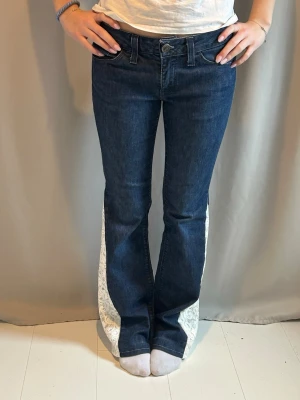 Mörkblå utsvängda jeans med spets - Unika mörkblå jeans med utsvängda ben och vita spetsinslag längs sidorna. Klassisk femficksmodell med låg midja och knappgylf. Perfekta för dig som vill sticka ut med en cool och personlig stil.🩵 innerbenslängden är 80cm och midjemåttet är 39cm rakt över🩵