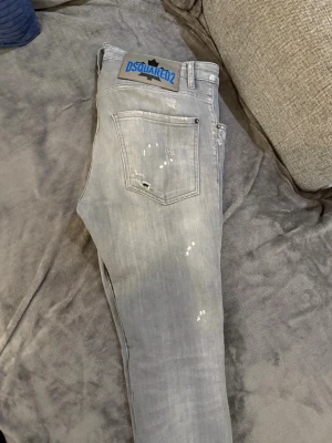 Ljusgrå Dsquared2 jeans - Säljer ett par ljusgrå jeans från Dsquared2 med coola slitningar och vita färgstänk. Jeansen har klassisk femficksmodell, rak passform och en stor Dsquared2-lapp bak i midjan. Tillverkade i bomull med en snygg tvättad look. Det är storlek m och s men det står 46 för det är dsquard storlek