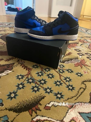 Air Jordan 1 Mid svart/blå  - Snygga Air Jordan 1 Mid sneakers i svart och blått läder med vita sulor och svarta skosnören. Klassisk siluett med ikonisk Jumpman-logga på plösen och Swoosh på sidan. Perfekta för dig som gillar streetwear och vill sticka ut.