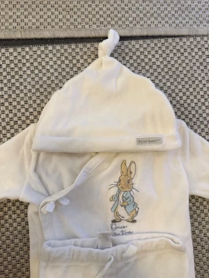 Vit set Peter Rabbit baby - Supersöt vit set för baby med broderad Peter Rabbit-figur och texten 'Once upon a time' framtill. Mjuk och gosig i frottéliknande material, knytband i midjan och huva med liten knut på toppen. Perfekt för mysiga morgnar hemma.