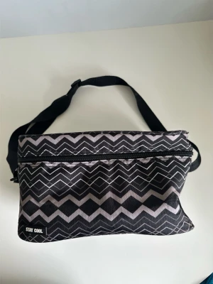 Svart Stay Cool kylväska med zigzag-mönster - Snygg kylväska från Stay Cool i svart med vitt zigzag-mönster. Väskan har justerbar axelrem och dragkedja upptill. Perfekt för picknicken, stranden eller som lunchväska. Slitstark vattenavvisande textil med vaddering och termisk funktion. Mått: 28x20x16cm. Fint skick!