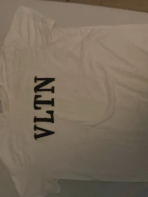 Vit VLTN t-shirt från Valentino - Säljer en klassisk vit t-shirt från Valentino med stor svart VLTN-logga framtill. T-shirten har rund hals och korta ärmar. Perfekt för dig som gillar stilrena och exklusiva plagg med tydlig logga. Märket blir kamouflage när den är i värme