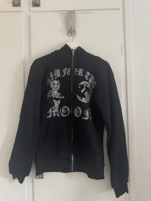 Svart hoodie Aim For The Moon fullzip  - Svart hoodie med dragkedja från Aim For The Moon. Framsidan har ett stort tryck med texten 'AIM FOR THE MOON' i glittriga stenar och motiv av en tecknad figur och måne.