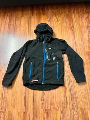 Svart vindjacka med blå dragkedjor - Snygg svart vindjacka från Team Sportia med blå dragkedjor och huva. Jackan har tre fickor med dragkedja, justerbar huva och sportig design. Perfekt för blåsiga dagar och outdoor-aktiviteter. Materialet känns lätt och smidigt.