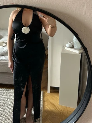 Svart långklänning med rosdetalk & slits - Snygg svart långklänning från Ginatricot med halterneck och öppen rygg. Klänningen har en djup v-ringning och ros framtill. Helt oanvänd med prislapp kvar✨🤍