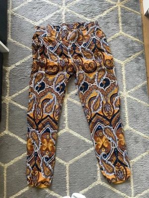 Mönstrade paisley-leggings i orange - Säljer ett par leggings med färgstarkt paisleymönster i orange, blått, vitt och brunt. De har elastisk midja och är tillverkade i ett mjukt, stretchigt material som sitter skönt. Perfekta för dig som vill sticka ut med din stil!